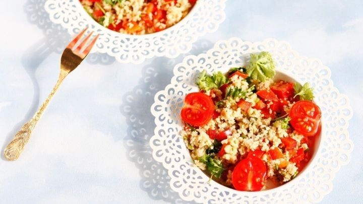 Zwei helle Schalen mit warmem Couscoussalat sind sch&ouml;n angerichtet, umgeben von einem hellblauen Hintergrund.