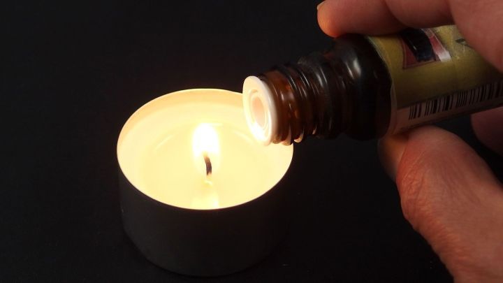 Ein Teelicht brennt, w&auml;hrend eine Hand Duft&ouml;l dar&uuml;bertr&auml;ufelt, um den Raum mit angenehmem Aroma zu f&uuml;llen.