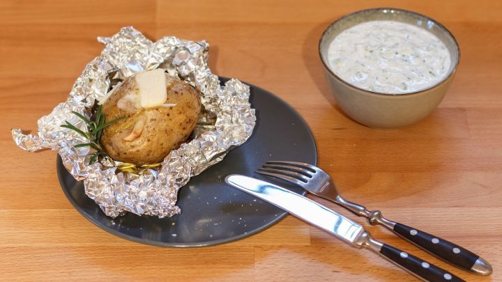 Eine Backkartoffel, in Alufolie gewickelt, liegt auf einem Teller neben einer Sch&uuml;ssel mit Kr&auml;uterquark.