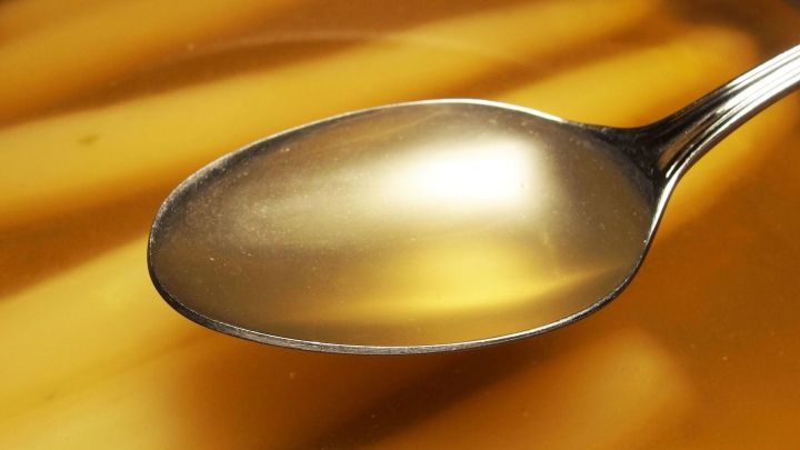 Eine silberne L&ouml;ffelspitze taucht ins klare Spargelkochwasser, das neben frisch gekochtem Spargel steht.