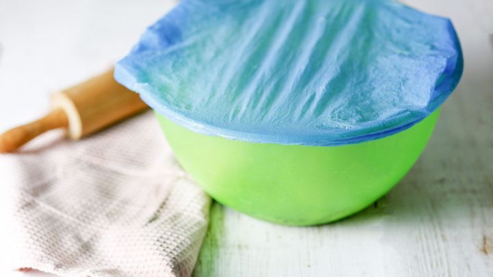 Eine gr&uuml;ne Sch&uuml;ssel mit Hefeteig, abgedeckt mit einer blauen Folie, steht auf einem Holztisch neben einem Nudelholz und einem Geschirrtuch.