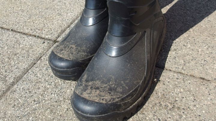 Schwarze Gummistiefel stehen auf einem grauen Gehweg und sind mit Schmutzflecken verunreinigt, bereit zum S&auml;ubern.
