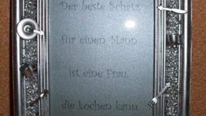 Ein Rahmen mit K&uuml;chenutensilien als Dekoration zeigt den Spruch: "Der beste Koch ... ist eine Frau, die kochen kann."
