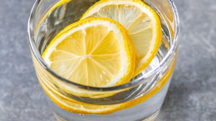 Ein Glas mit klarem Wasser und Zitronenscheiben schwimmt, umgeben von einer grauen Oberfl&auml;che, die frisch und einladend wirkt.