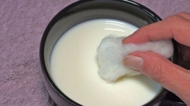 Eine Hand taucht einen Wattepad in Buttermilch, um das Gesicht abzuschminken, neben einer weichen, lila Unterlage.