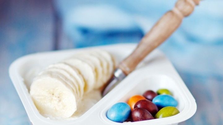 Ein leeres Joghurtbeh&auml;lter mit Bananenscheiben und bunten Schokolinsen steht auf einem rustikale Holzuntergrund.