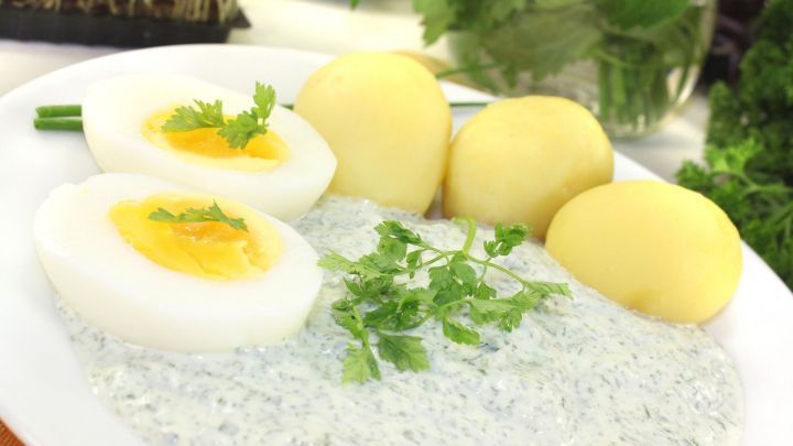 Ein Teller mit Kasseler Gr&uuml;ne So&szlig;e, gekochten Eiern und Kartoffeln, umgeben von frischen Kr&auml;utern.