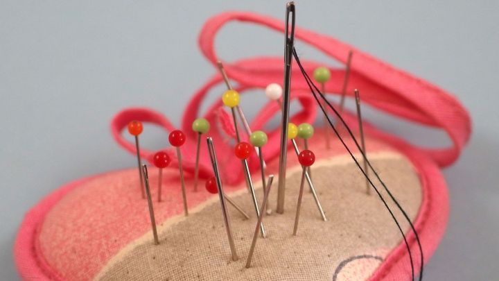 Eine Nadel mit Faden erhebt sich aus einem Nadelkissen, w&auml;hrend mehrere Stecknadeln darin stecken.