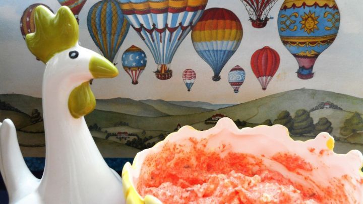 Eine wei&szlig;e H&uuml;hnerfigur steht neben einer rosa Lachsbutter in einer bunten Sch&uuml;ssel, vor einer Wand mit Hei&szlig;luftballons.