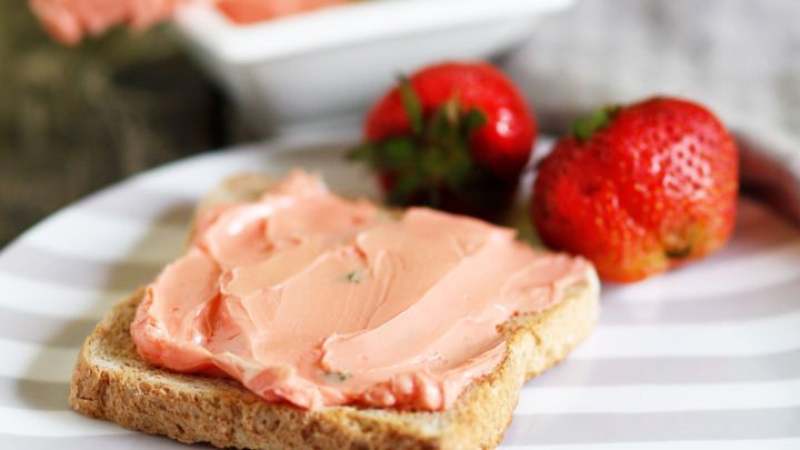 Eine Scheibe Vollkornbrot ist mit Erdbeerbutter bestrichen, w&auml;hrend ein Dip mit frischer Minze im Hintergrund ist. Frische Erdbeeren liegen daneben.