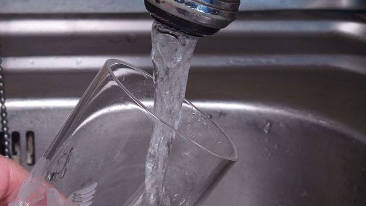 Ein Glas wird unter einem Wasserhahn gef&uuml;llt, w&auml;hrend frisches Wasser flie&szlig;t, im Kontext einer K&uuml;chensp&uuml;le.