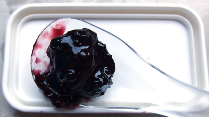 Dunkle Marmelade wird mit einem L&ouml;ffel aus einer wei&szlig;en Schale entnommen. Der Hintergrund ist unauff&auml;llig.