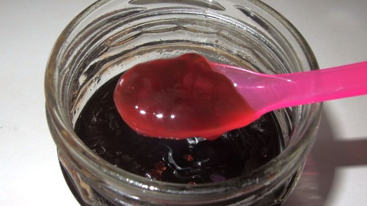 Eine pinke L&ouml;ffelspitze hebt Marmelade aus einem Glas, die etwas zu d&uuml;nn wirkt, auf einem hellen Tisch.