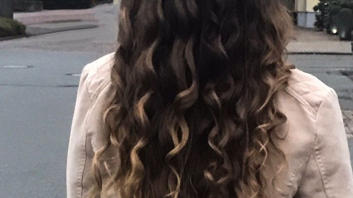 Lange, gl&auml;nzende Locken fallen &uuml;ber die Schultern einer Person, die mit dem R&uuml;cken zur Kamera auf einer Stra&szlig;e steht.