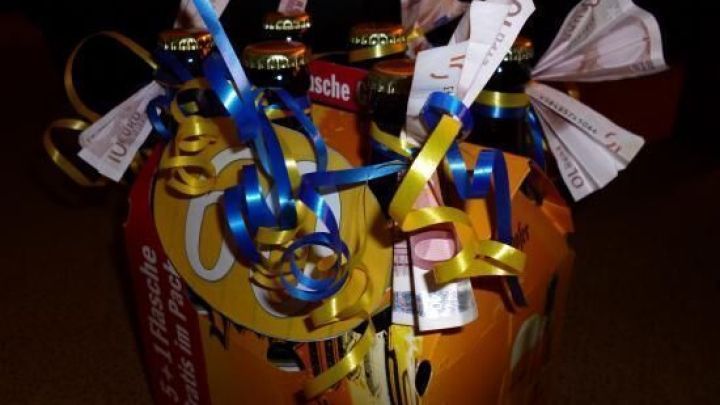 Ein Sechserpack Bierflaschen, dekoriert mit bunten B&auml;ndern und 10-Euro-Geldscheinen, im Halbkarton auf Teppichboden.