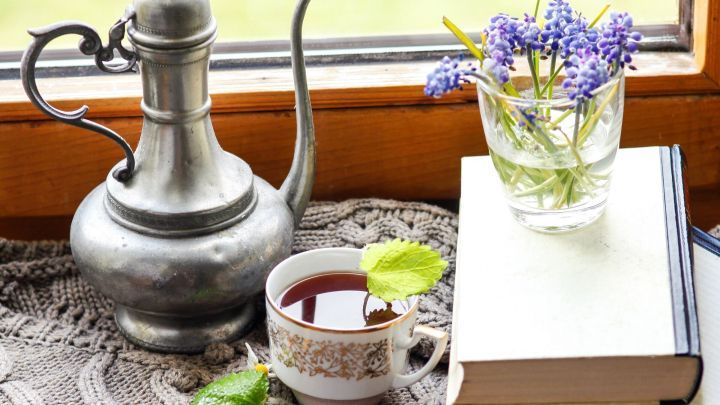 Eine Teetasse steht auf einem Tisch, w&auml;hrend eine Teekanne und Blumen in einem Glas neben einem offenen Buch arrangiert sind.