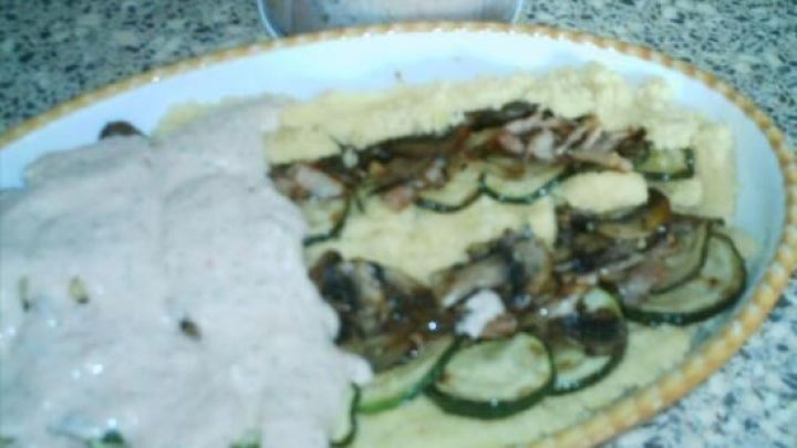 Ein Auflauf aus Polenta liegt auf einem Teller, garniert mit Zucchini und einer cremigen Sauce, in einer hellen Umgebung.