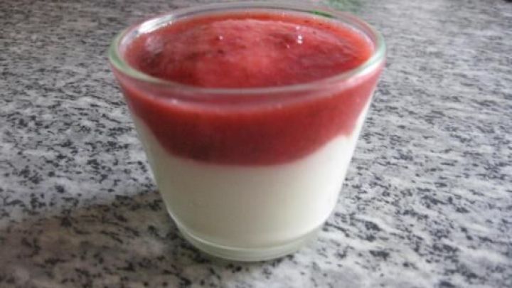 Ein Dessert mit wei&szlig;er Schokolade und einer fruchtigen Erdbeerso&szlig;e in einem Glas auf einer grauen Arbeitsplatte.