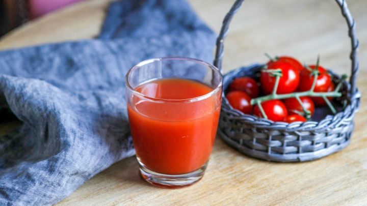 Ein Glas Selbstgemachter Tomatensaft steht auf einem Tisch neben einem Korb voller frischer Tomaten, umgeben von einem eleganten, gepolsterten Stuhl.