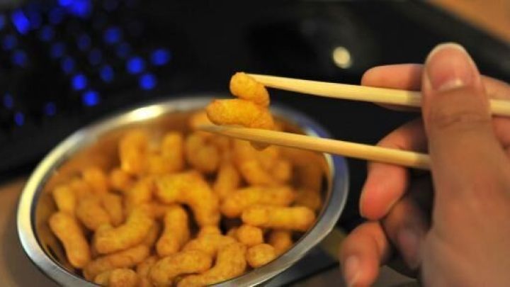 Eine Hand h&auml;lt Essst&auml;bchen, die eine Erdnussflip-Locke aus einer Sch&uuml;ssel aufnehmen, w&auml;hrend ein Computer im Hintergrund sichtbar ist.