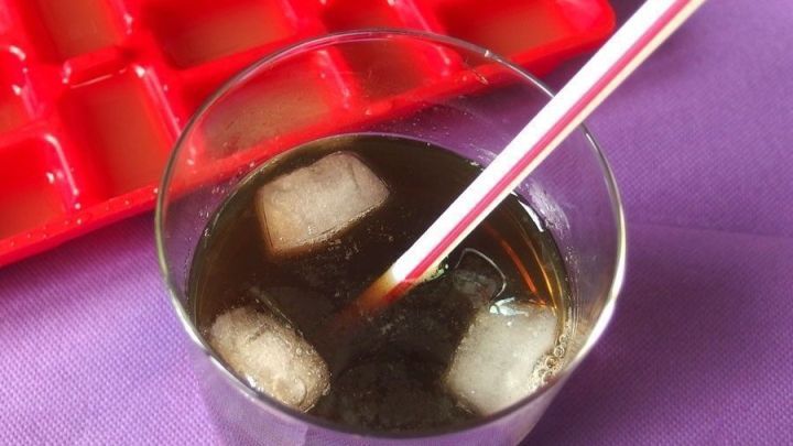 Ein Glas selbstgemachter Eistee mit Eisw&uuml;rfeln und einem Strohhalm steht auf einem lila Tisch, w&auml;hrend im Hintergrund eine rote Eisw&uuml;rfelform sichtbar ist.