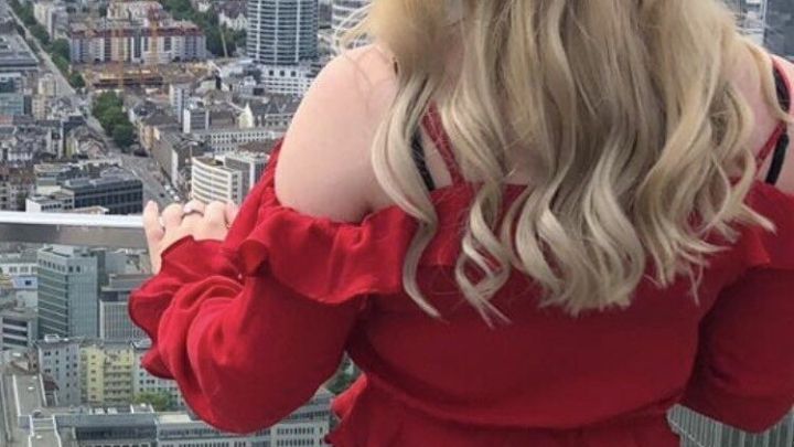 Eine Frau mit lockigen Haaren steht am Rande eines Hochhauses und blickt auf die Stadtlandschaft.