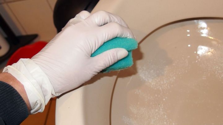 Eine Hand mit einem Einmal-Gummihandschuh h&auml;lt einen gr&uuml;nen Schwamm, w&auml;hrend sie eine Toilette reinigt.