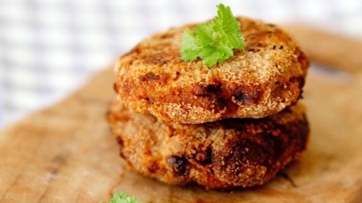 Zwei goldbraune Frikadellen liegen auf einem Holzbrett, garniert mit frischem Koriander, in einer gem&uuml;tlichen K&uuml;chenszene.