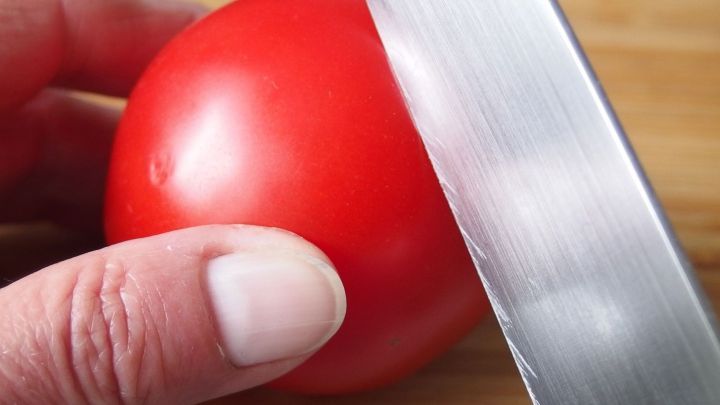 Eine Hand h&auml;lt eine Tomate, w&auml;hrend ein Messer sie gerade schneidet, auf einer Holzfl&auml;che in einer K&uuml;chenumgebung.