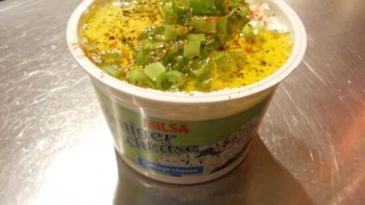 Ein Becher H&uuml;ttenk&auml;se steht auf einer K&uuml;chentheke, garniert mit gr&uuml;nen Zwiebeln, umgeben von einer modernen K&uuml;chenumgebung.