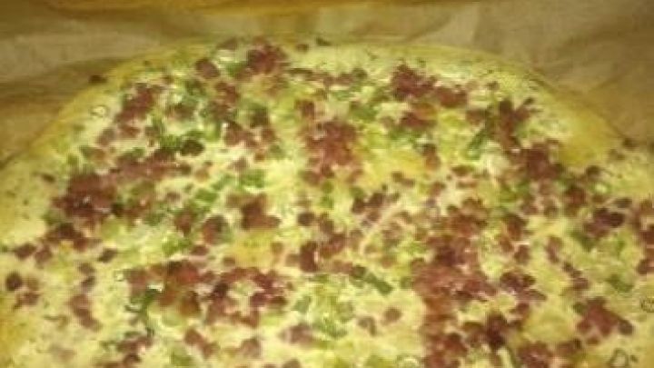 Ein runder Flammkuchen mit Hefeteig, belegt mit Speckw&uuml;rfeln und Fr&uuml;hlingszwiebeln, ruht auf Backpapier auf Holzoberfl&auml;che.