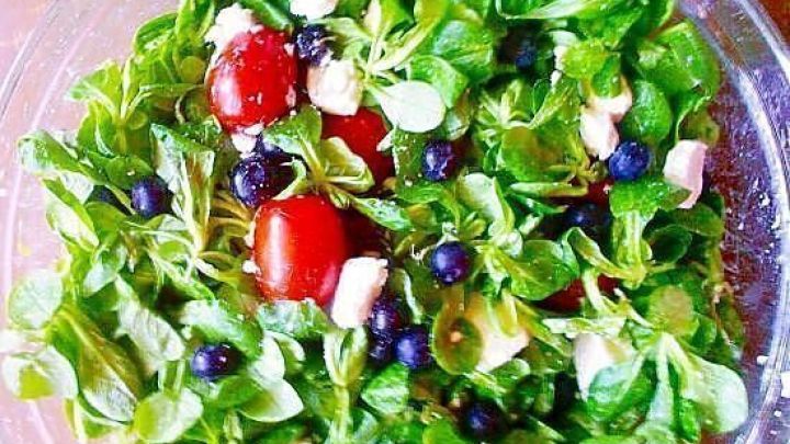 Feldsalat mischt sich mit frischen Blaubeeren und roten Tomaten in einer klaren Sch&uuml;ssel auf einem Holztisch.