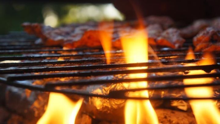 Flammen lodern unter einem Grillrost, auf dem Fleischst&uuml;cke liegen, im Hintergrund verschwommenes Gr&uuml;n.
