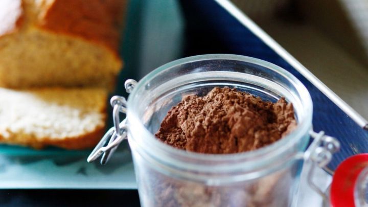 Eine Glasdose mit Kakaopulver steht auf einem Tisch, daneben liegt frisch gebackenes Brot in einem rustikalen Umfeld.