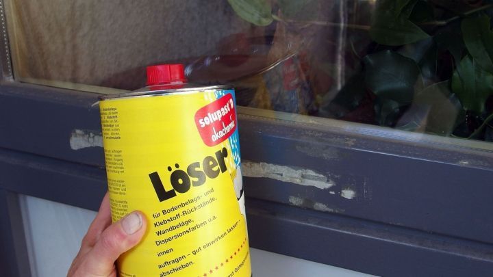 Ein Hand h&auml;lt eine gelbe Dose mit "L&ouml;ser" neben einem Fensterrahmen, der Klebereste aufweist, in einem hellen Innenraum.