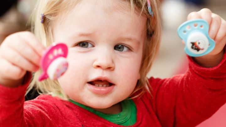 Ein Kleinkind h&auml;lt zwei Schnuller, einen rosa und einen blauen, in den H&auml;nden und schaut neugierig in einer h&auml;uslichen Umgebung.