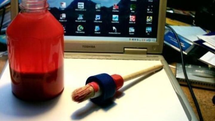 Ein Pinsel liegt neben einer roten Flasche auf einem ge&ouml;ffneten Laptop mit Desktop-Icons im Hintergrund.