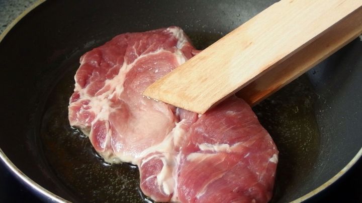 Zwei rohe St&uuml;cke Fleisch werden in einer Pfanne braten, w&auml;hrend ein Holzspatel das Fleisch umdreht.