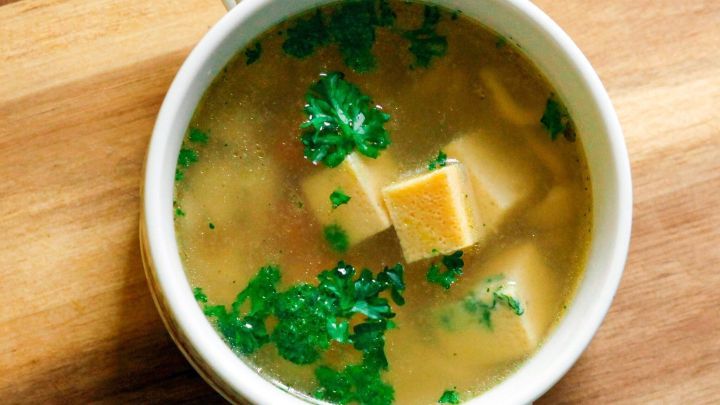 Eine helle Suppe mit gr&uuml;ner Petersilie und K&auml;sew&uuml;rfeln wird in einer wei&szlig;en Sch&uuml;ssel auf einem Holzbrett serviert.