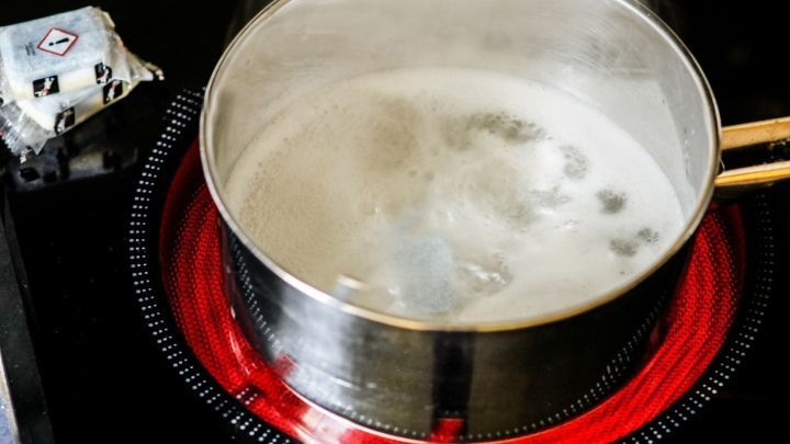 Ein Kochtopf mit kochendem Wasser steht auf einem hei&szlig;en Herd, w&auml;hrend Dampf aufsteigt und etwas daneben liegt.