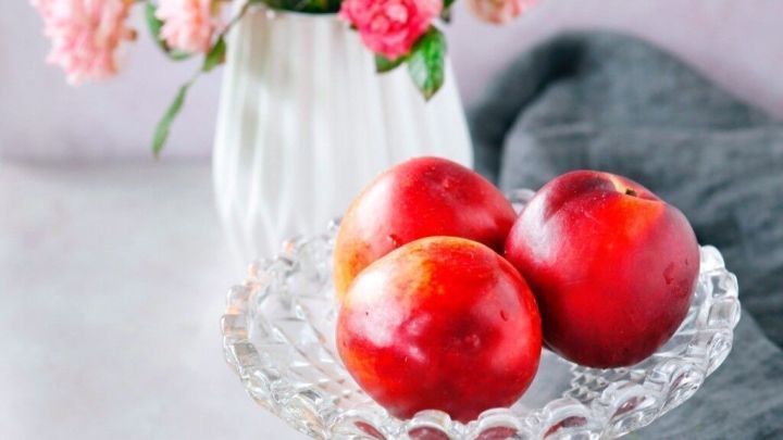 Reife Nektarinen liegen auf einem eleganten Stand im Vordergrund, w&auml;hrend eine Blume im Hintergrund eine sch&ouml;ne Atmosph&auml;re schafft.