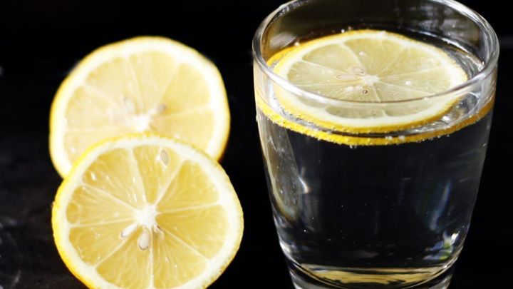 Ein Glas Mineralwasser mit Zitronenscheiben erfrischt in einem dunklen Umfeld, w&auml;hrend zwei halbierte Zitronen daneben liegen.