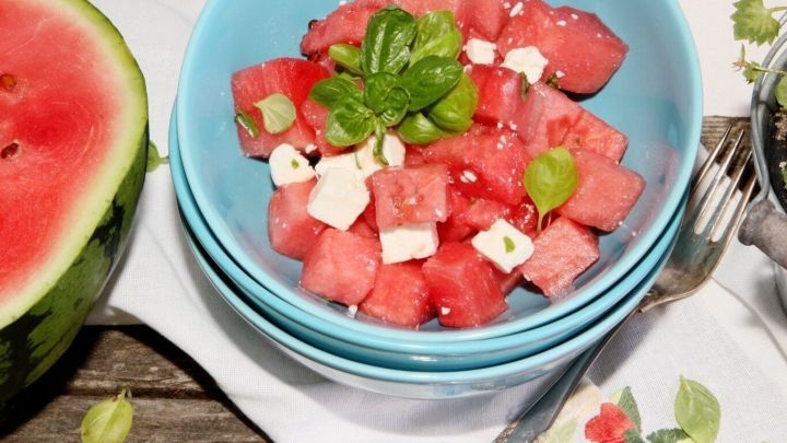 Eine Sch&uuml;ssel mit Wassermelone und Feta ist dekorativ angerichtet, umgeben von frischen Kr&auml;utern auf einem Tisch.