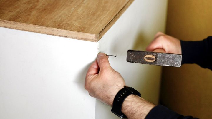 Ein Handwerker schl&auml;gt einen kleinen Nagel in ein IKEA-Regal, w&auml;hrend er mit einem Hammer arbeitet und den Regalkorpus repariert.