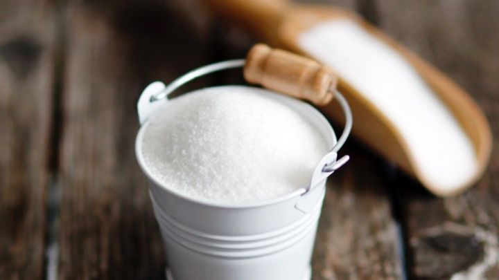 Ein kleiner Eimer mit Zucker steht auf einem Holztisch, w&auml;hrend ein Holzl&ouml;ffel daneben liegt. Der Zucker k&ouml;nnte gegen Schluckauf helfen.