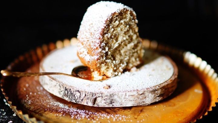 Ein Nusskuchen mit Puderzucker best&auml;ubt liegt auf einem rustikalen Holzbrett, umgeben von einem gl&auml;nzenden Teller.