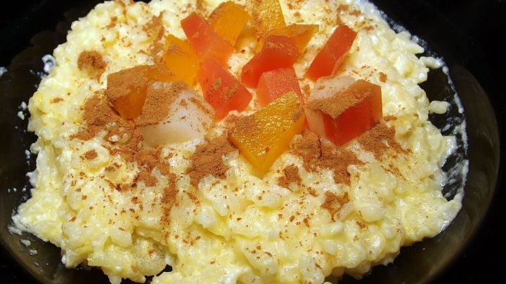 Die Sch&uuml;ssel zeigt cremigen Milchreis, garniert mit bunten Fruchtst&uuml;cken und Zimt, auf einem dunklen Teller serviert.