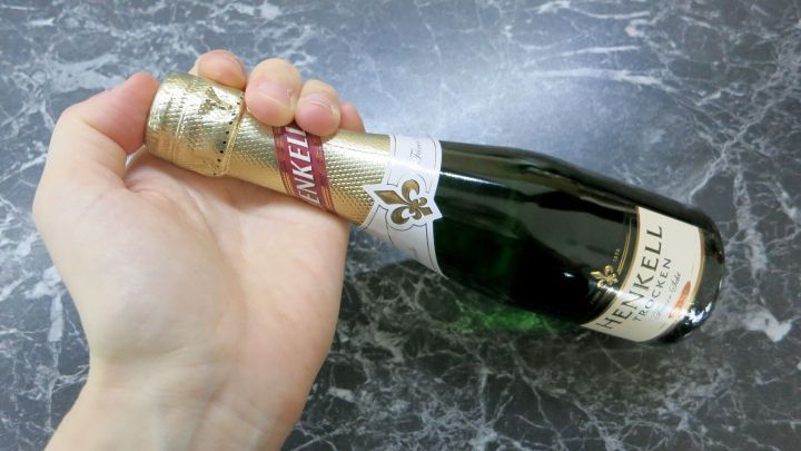 Eine Hand h&auml;lt eine Sektflasche mit HENKELL TROCKEN-Label auf einem dunklen Marmortisch.