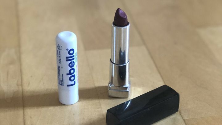 Ein Lippenstift und ein Lippenpflegestift liegen auf einem Holztisch, umgeben von weiteren Kosmetikprodukten.