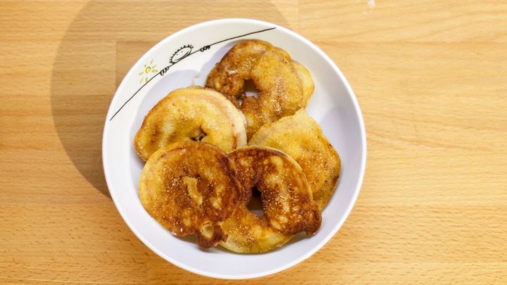 In einer wei&szlig;en Schale liegen goldene, frittierte Apfelk&uuml;chlein auf einem Holzuntergrund, bereit zum Genie&szlig;en.